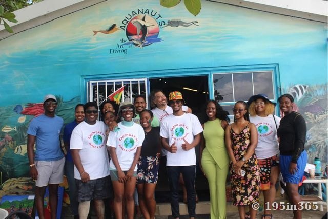 A large group of young people, many in white Earth Day ’24 “Make a Change” T-shirts, smiling outside a dive shop painted with an underwater mural; one holds the flag of Grenada.
