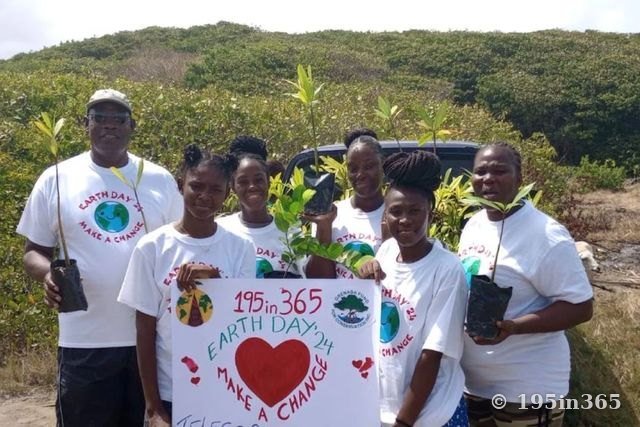 Six people in Earth Day ’24 T-shirts hold potted mangrove seedlings and a “195in365 Earth Day ’24 Make a Change” sign in front of coastal scrubland.