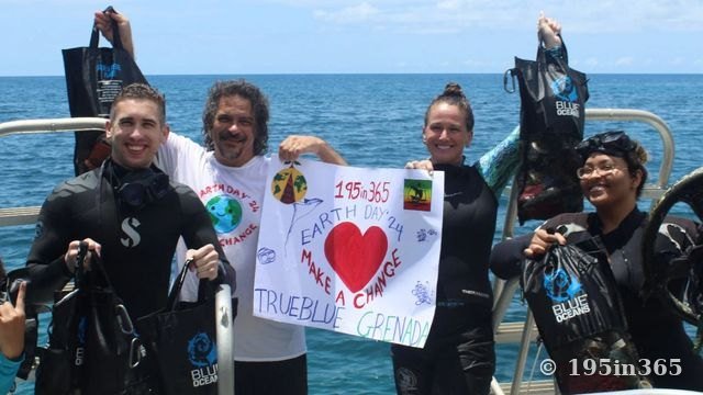 Divers in wetsuits on a boat hold up mesh bags of rubbish and a hand-painted “195in365 Earth Day ’24 Make a Change, TrueBlue Grenada” banner, with open sea behind them.
