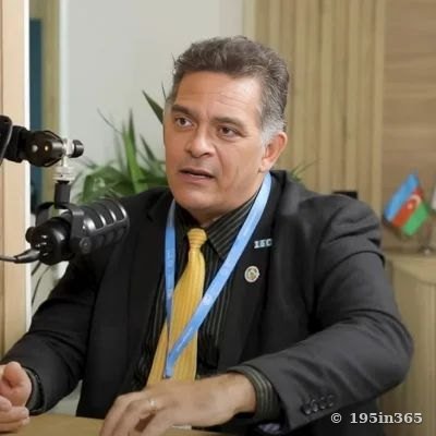 A man in a suit and a conference lanyard speaks into a studio microphone, with a flag on the table behind him.