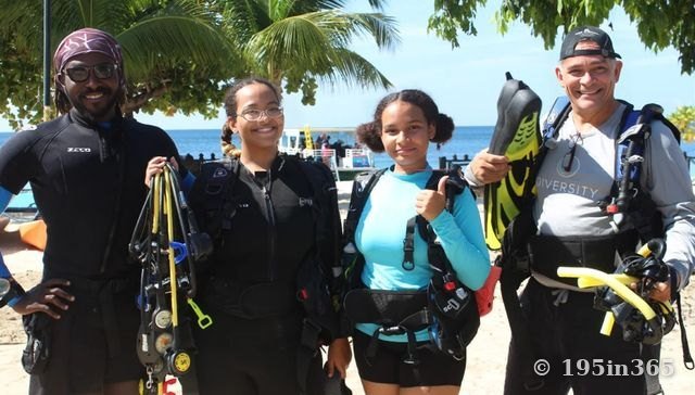 Four divers of different ages stand on a beach holding fins and dive equipment, smiling, with palm trees and a dive boat behind them.