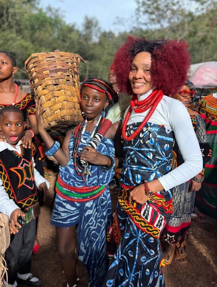 A smiling woman in a blue and black traditional outfit with red coral beads stands beside a girl carrying a woven basket, among people in colourful traditional dress at an outdoor gathering.