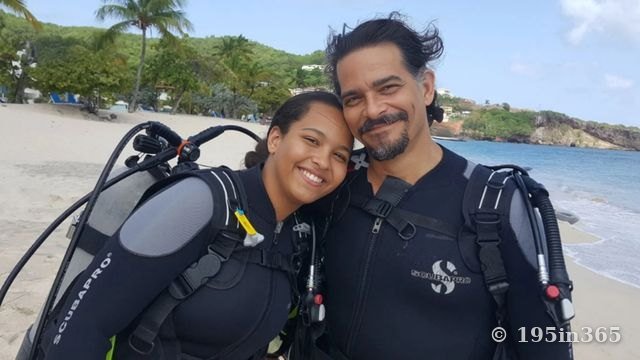 Una joven sonriente y un hombre, con trajes de neopreno y equipo de buceo, posan mejilla con mejilla en una playa soleada, con palmeras y el mar detrás.