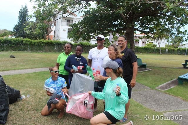 Siete voluntarios se reúnen bajo un gran árbol en un parque, con botellas de plástico y bolsas de basura recogida, riendo juntos.